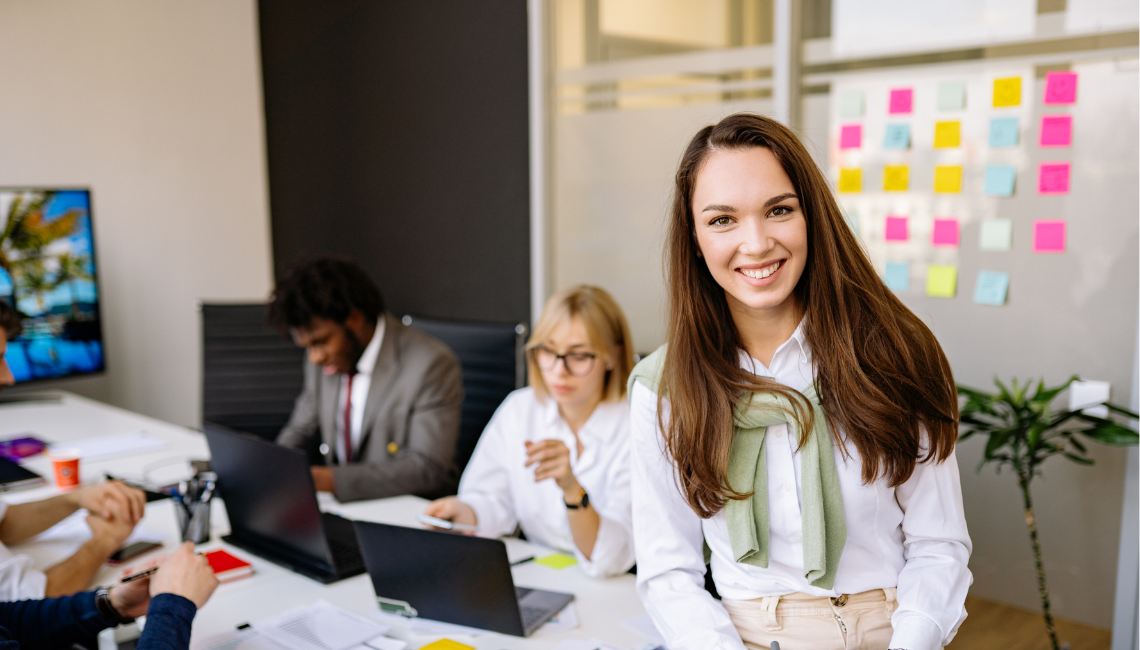 A smiling woman stands at the front of a team meeting in a modern office while colleagues work around a table.