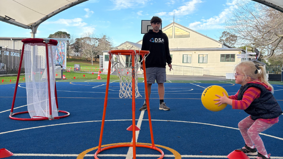 Young child holding a yellow ball on an outdoor court, preparing to play while an instructor stands beside adapted sports hoops.