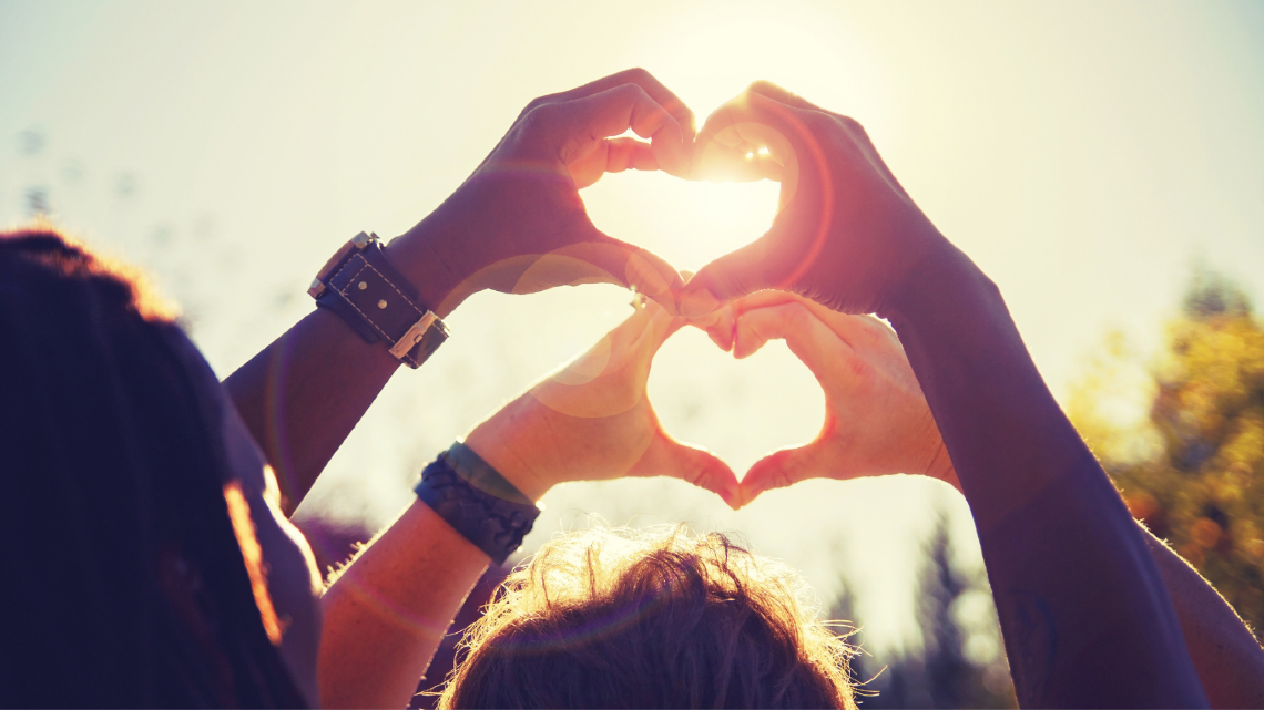 Two people form heart shapes with their hands in bright sunlight.