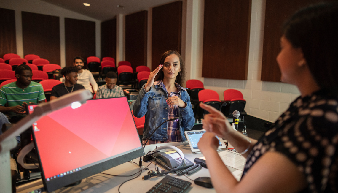 Woman signs at the front of a classroom while another tutor responds, with students watching during an NZSL lesson.