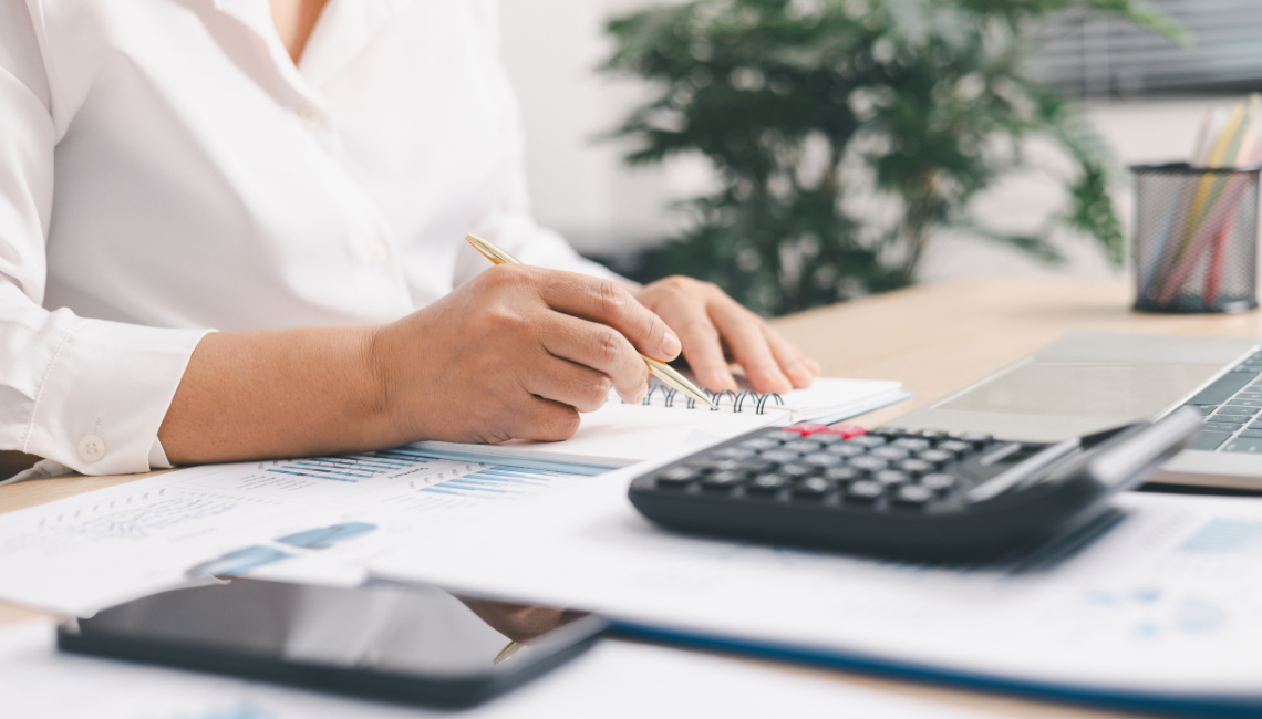 A person writes in a notebook beside a laptop, calculator, and printed financial documents on a desk.