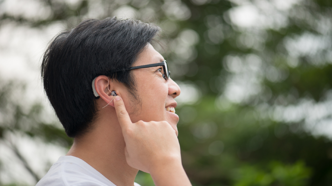 Person outdoors touching a hearing aid behind their ear.Person outdoors touching a hearing aid behind their ear.