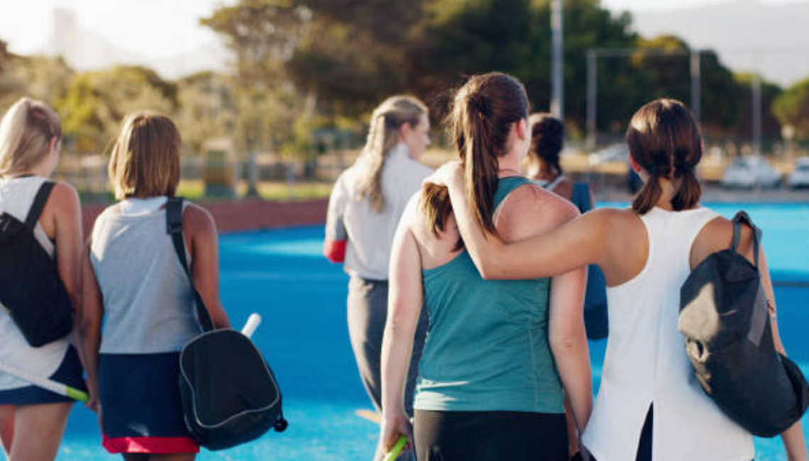 Girls in sportswear walk onto an outdoor court together, with one player’s arm around another in a supportive team moment.