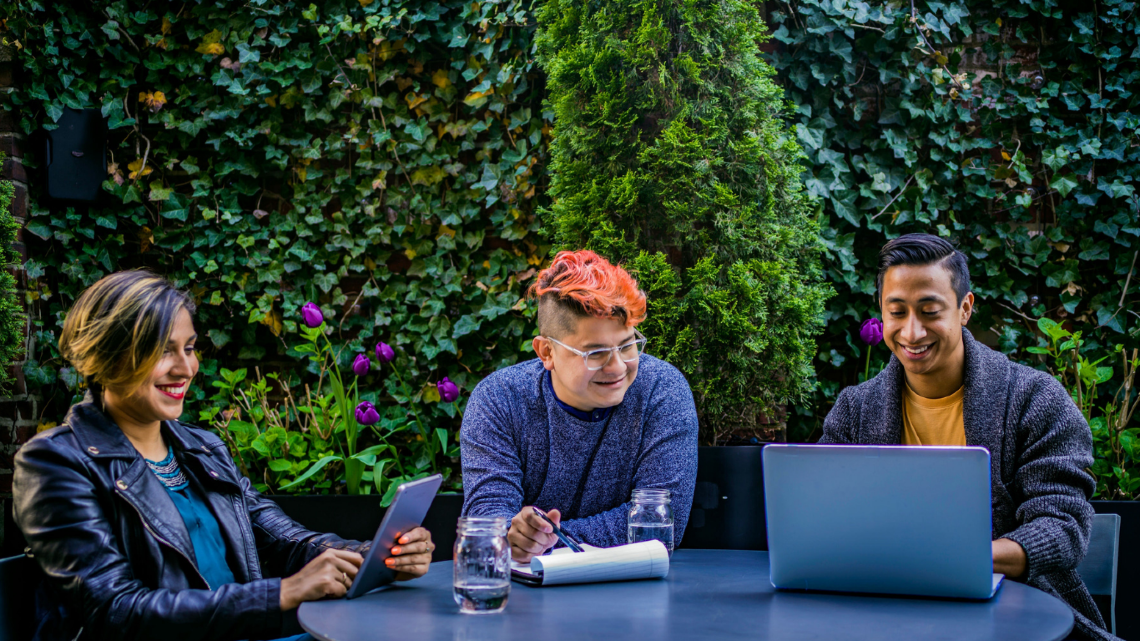 Three adults using laptops and a tablet together at an outdoor table.