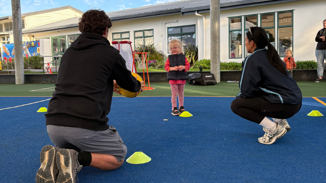 Two adults support a young child during an outdoor activity at an early learning centre.