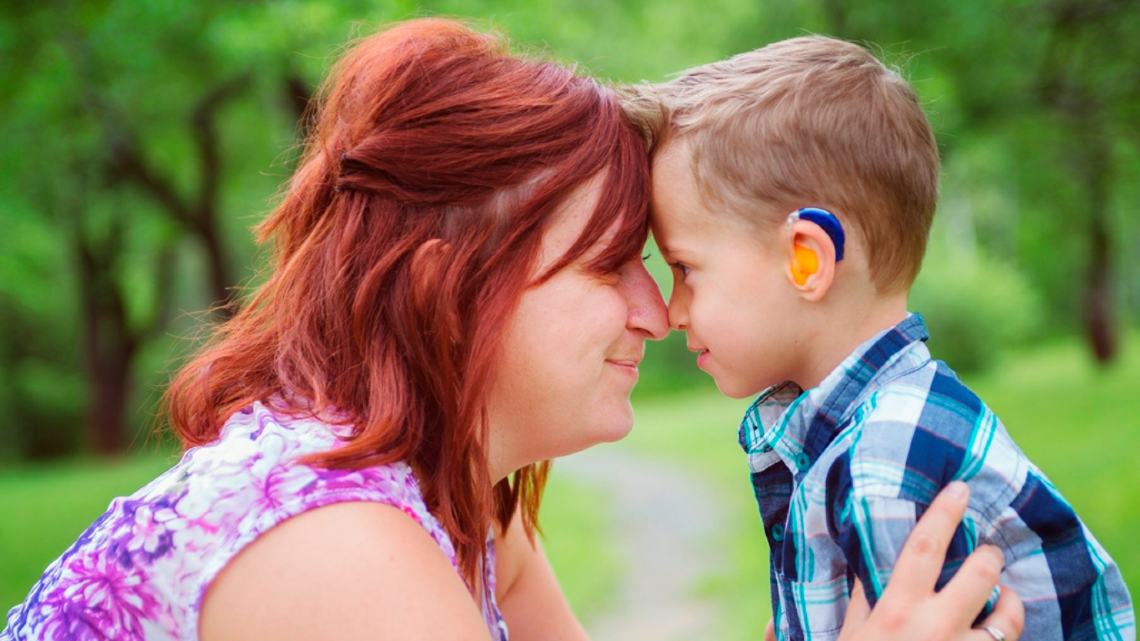 Woman and young boy with a hearing aid touch foreheads outdoors, showing close family connection and support.