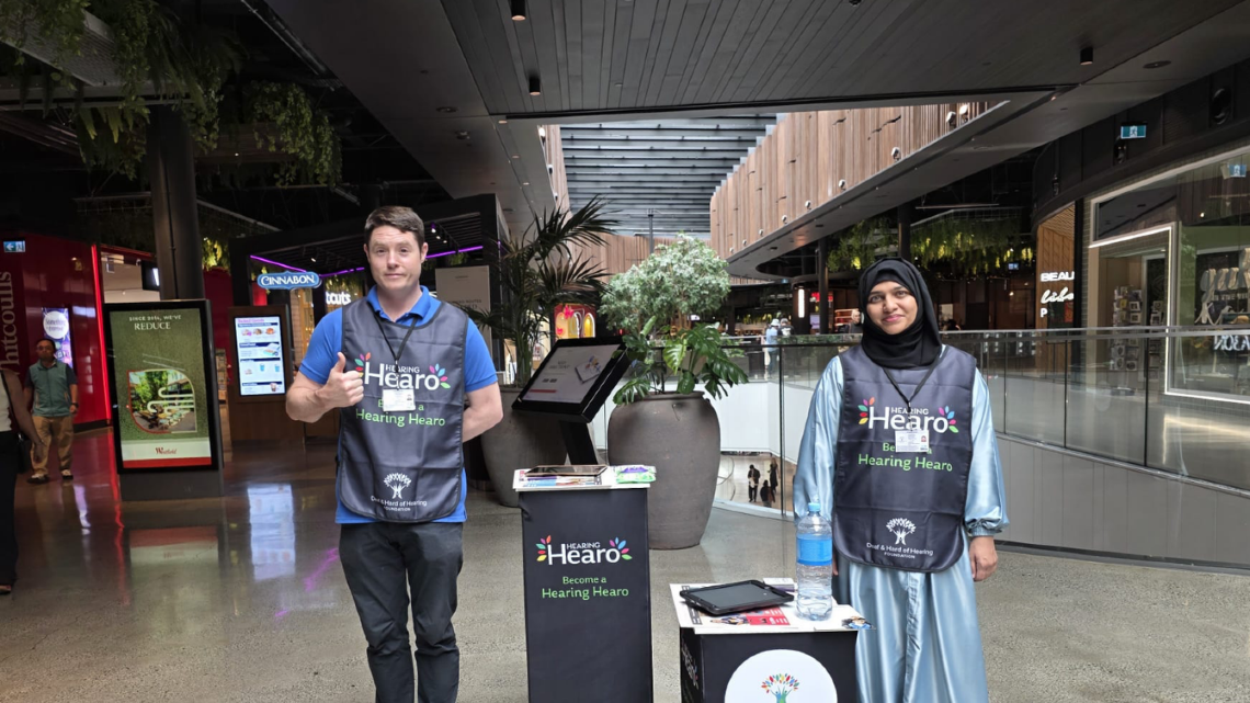 Two Face-to-Face fundraisers at a Hearing Hearo information stand in a shopping centre, inviting people to support the Deaf & Hard of Hearing Foundation.