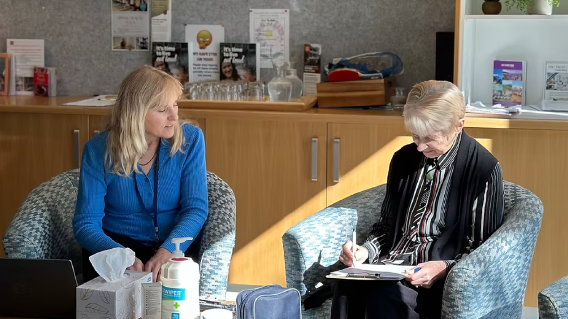 Staff member helping older woman fill out survey form seated at a retirement village outreach event.