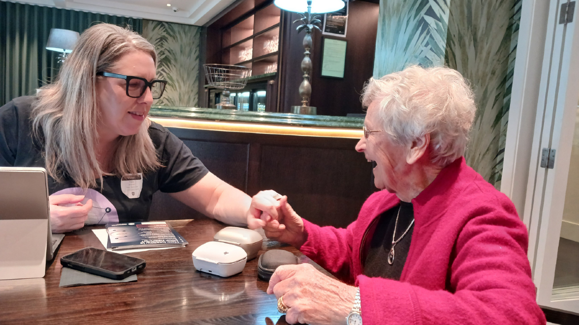 Staff member helping an older woman handle a hearing device during a retirement village visit.