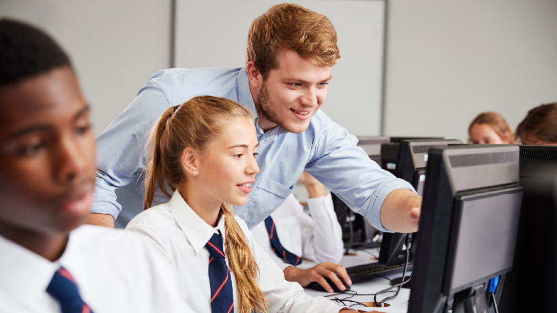 Teacher helping a student at a computer in a classroom.