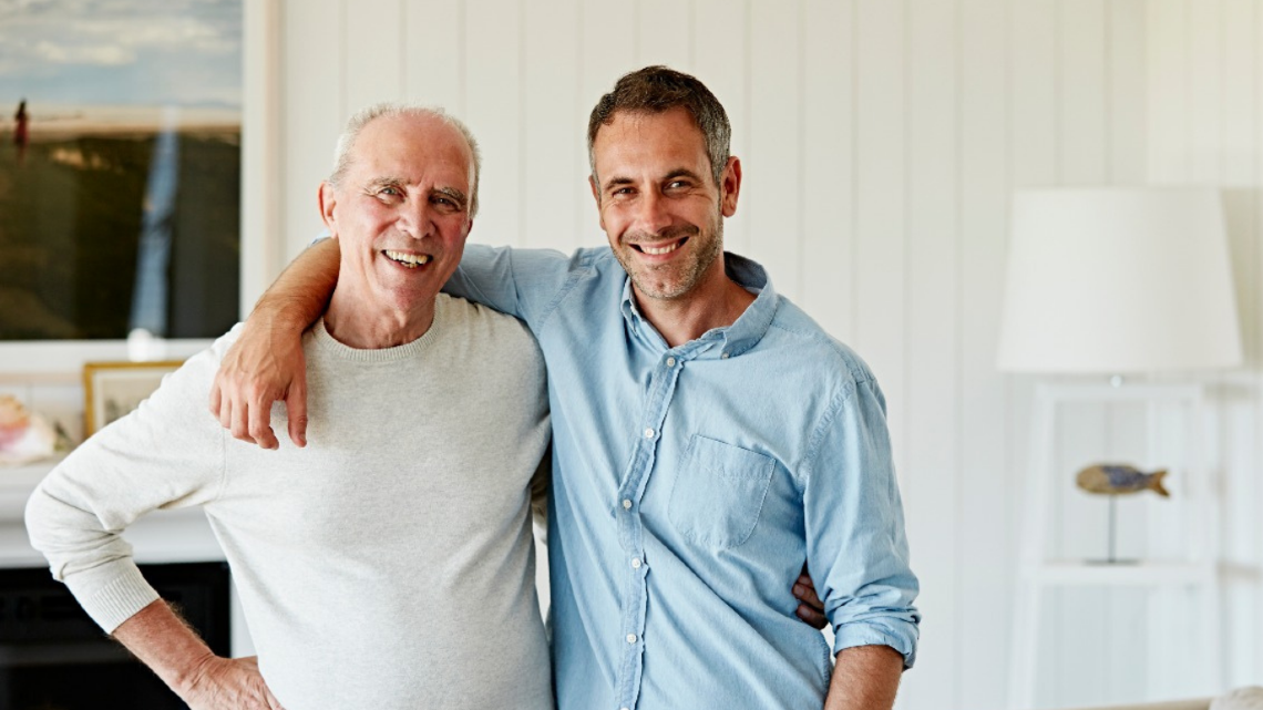 Two men smiling with their arms around each other at home, showing care, connection, and shared support.