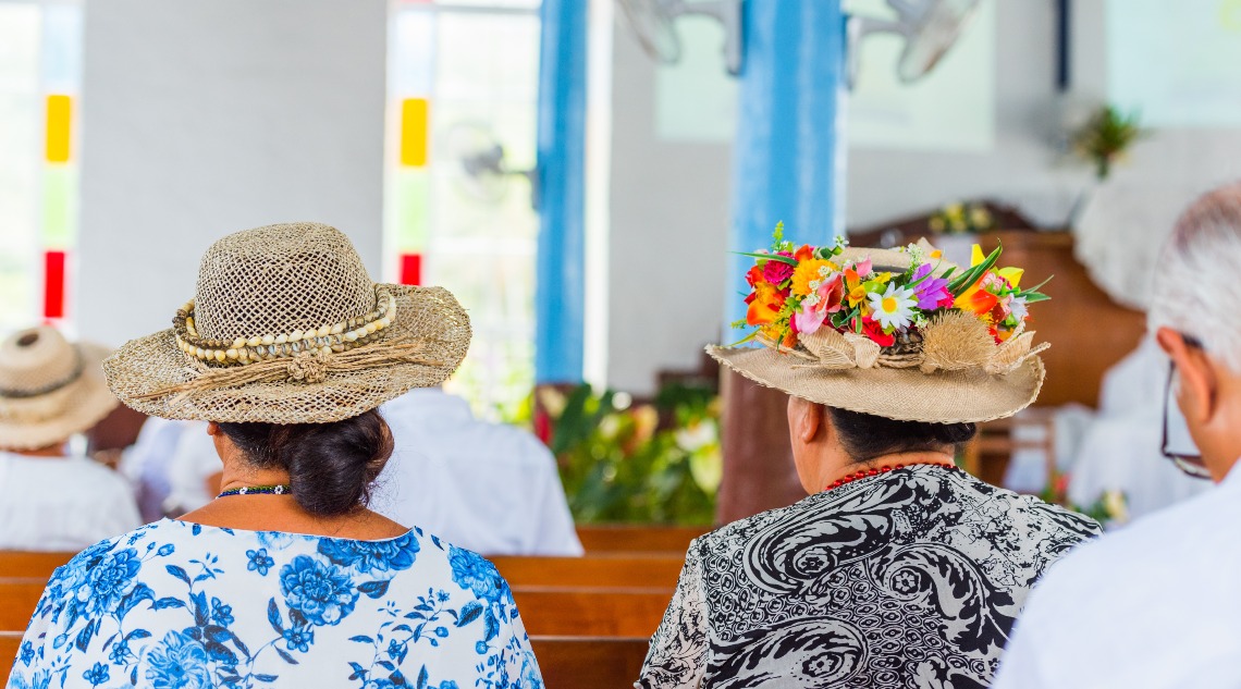 People seated in a church wearing decorated hats, reflecting Pasifika cultural tradition and community gathering.