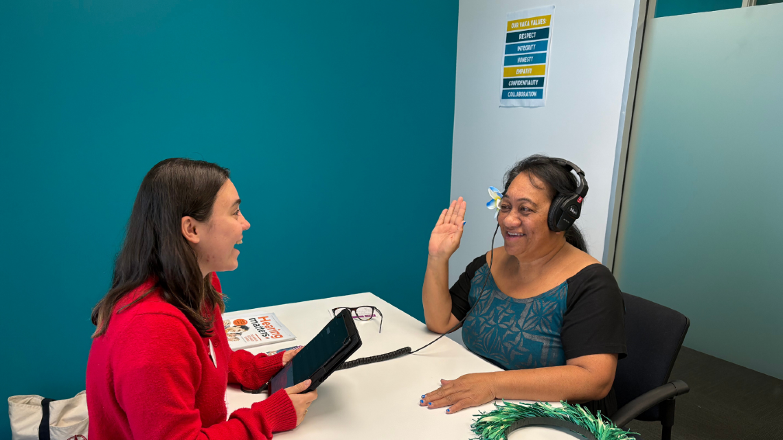 Two women talk across a table during a hearing support session, with one wearing headphones and smiling as they communicate.