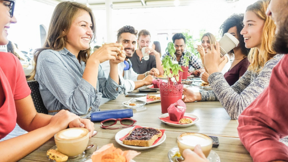 Group of people chatting and laughing around a café table, showing community connection and shared support.
