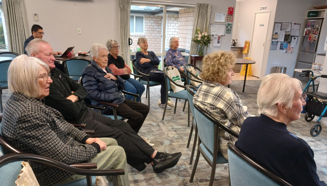 Older adults sit in a community room listening to a speaker during an information session.