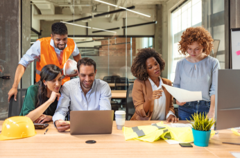Five staff members gathered around a laptop and paperwork, planning workplace NZSL support in a collaborative office setting.