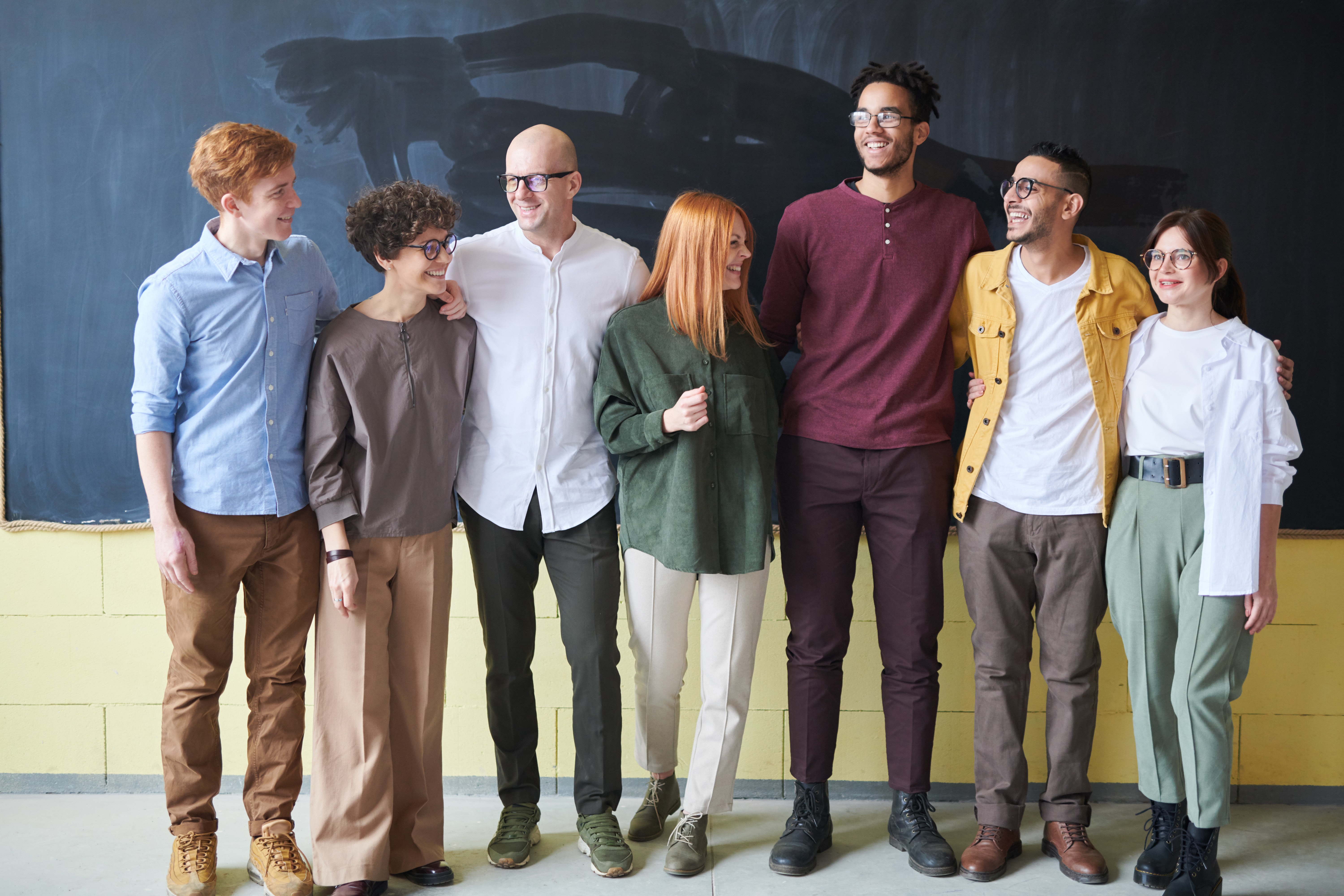 A diverse group of adults stand together smiling with their arms around each other, representing inclusion and community.