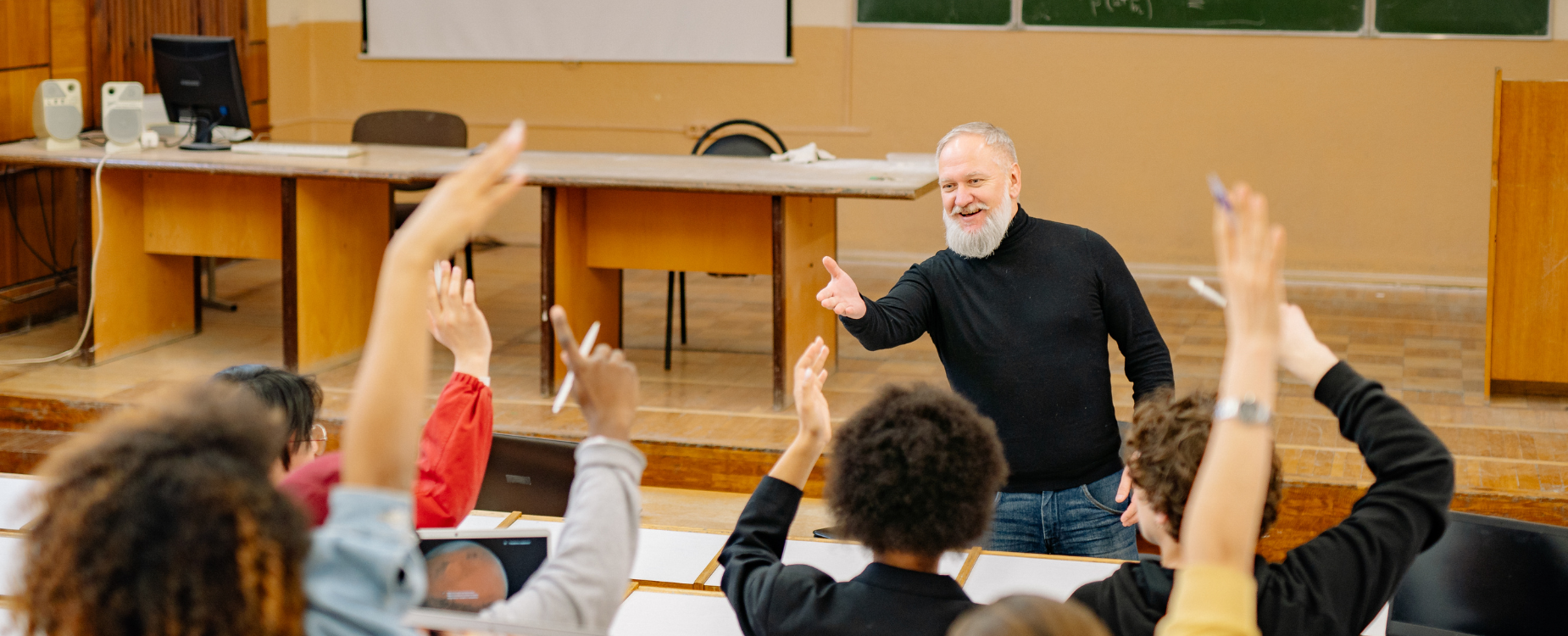 Lecturer smiling and speaking to a classroom of students with raised hands during a group discussion.