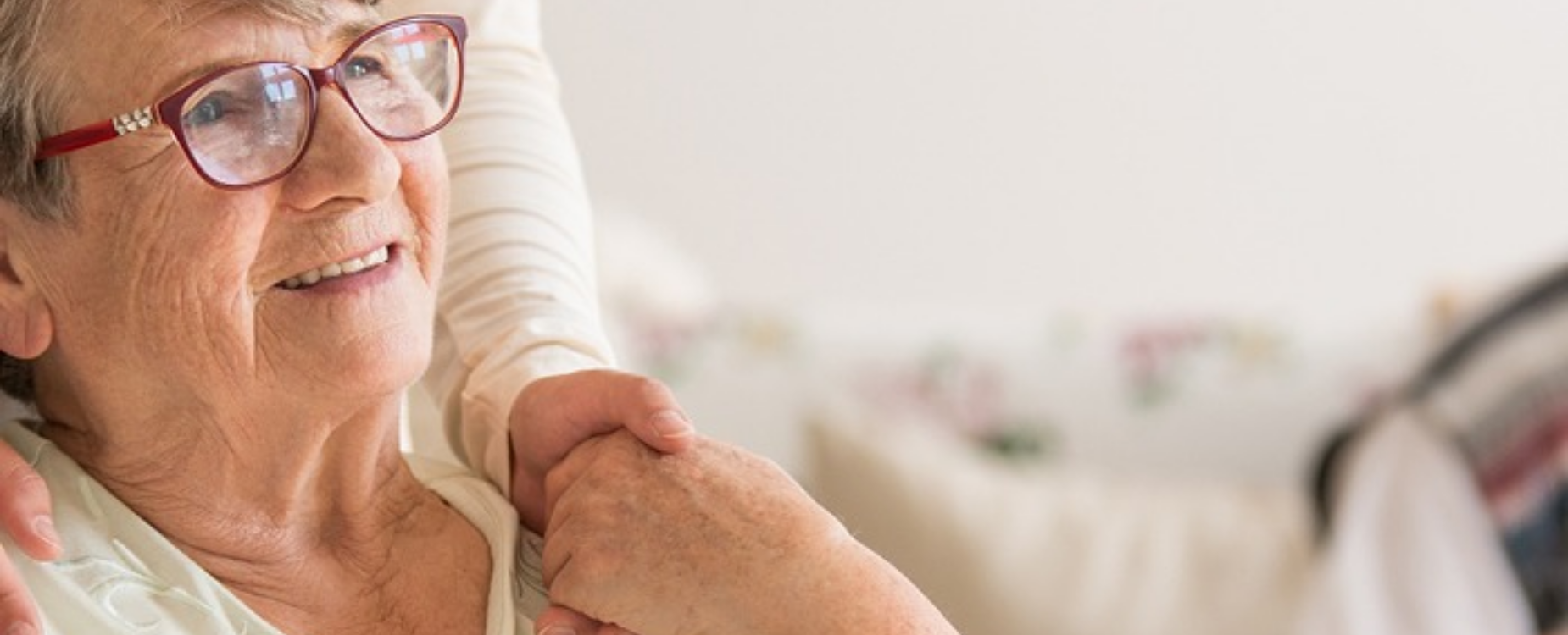 Older woman smiling while someone rests a hand on her shoulder, showing care, comfort, and personal support.