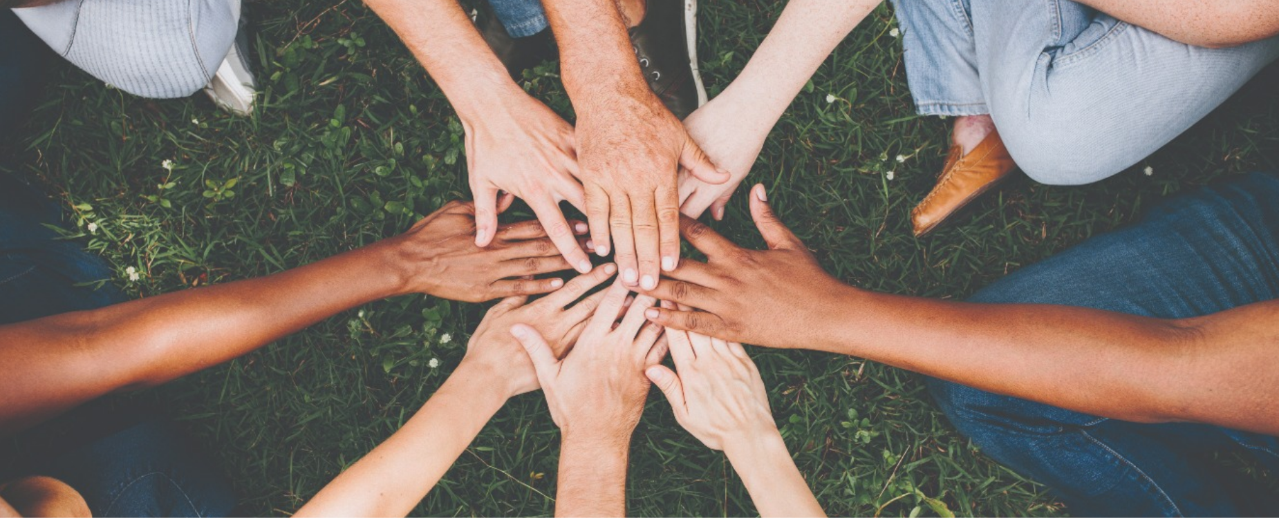 Many hands stacked together in a circle over grass, representing community connection and support during Hearing Awareness Month.