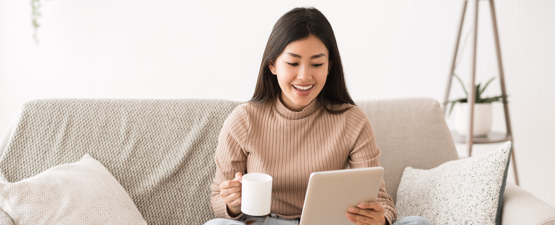 Woman smiling at a tablet while sitting on a sofa with a mug, suggesting easy access to hearing health information from home.