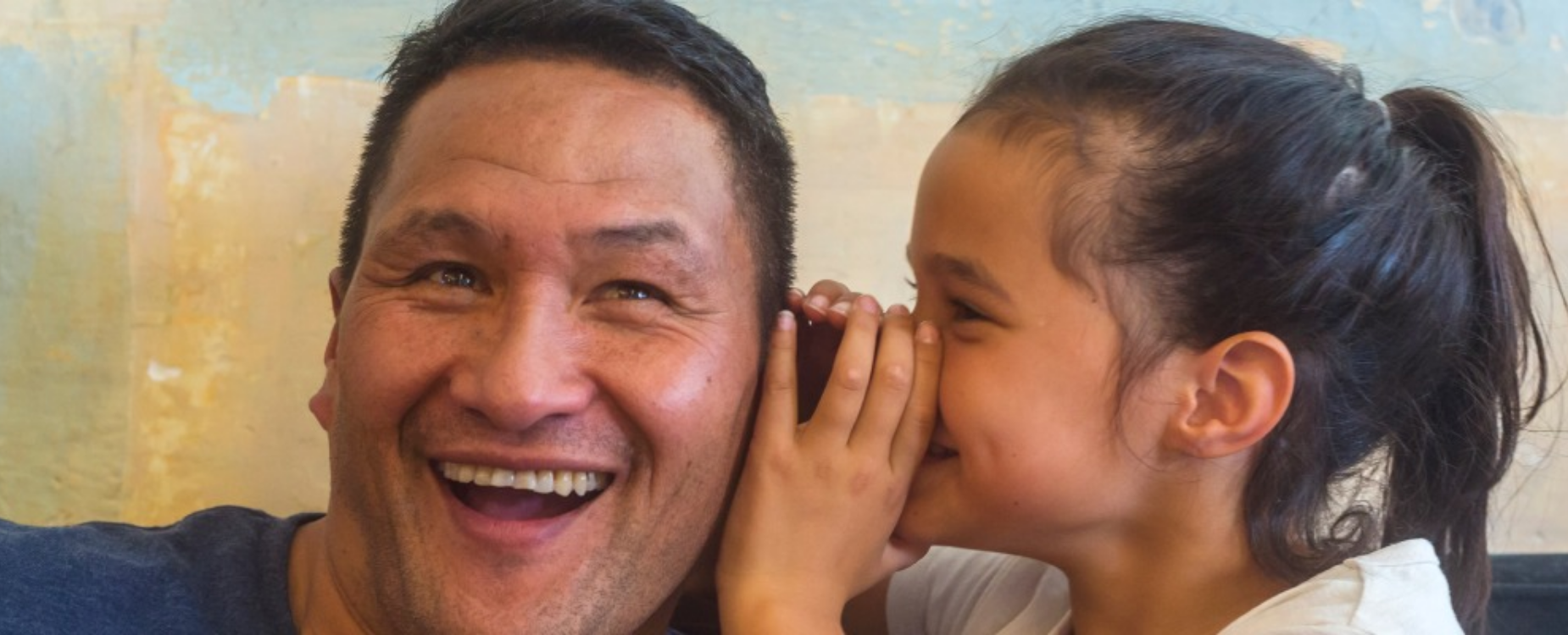 Young girl whispers into a smiling man&rsquo;s ear, sharing a playful moment together.