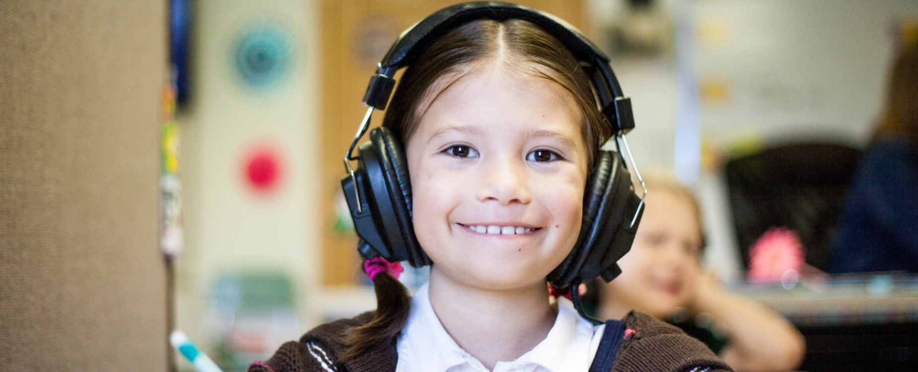 A child wearing headphones and smiling with the classroom in the background.