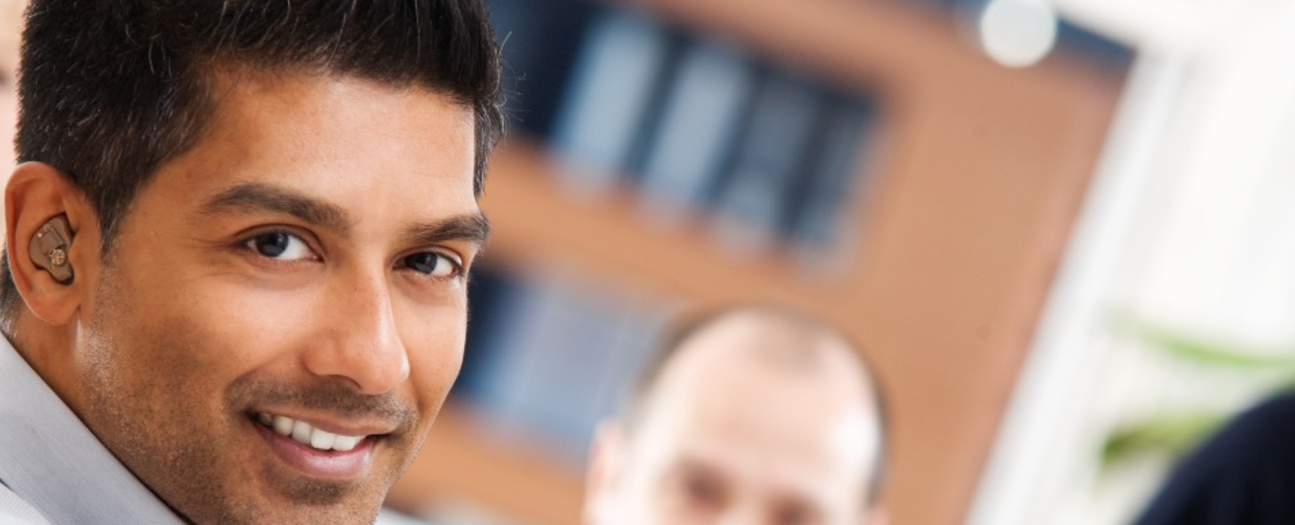 Man wearing a hearing aid smiling toward the camera, with other people blurred in the background on a media enquiries page.