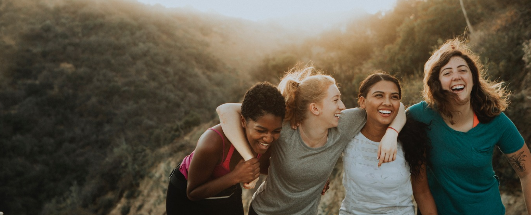 Four women laugh with their arms around each other on a hillside trail, sharing a joyful moment outdoors.