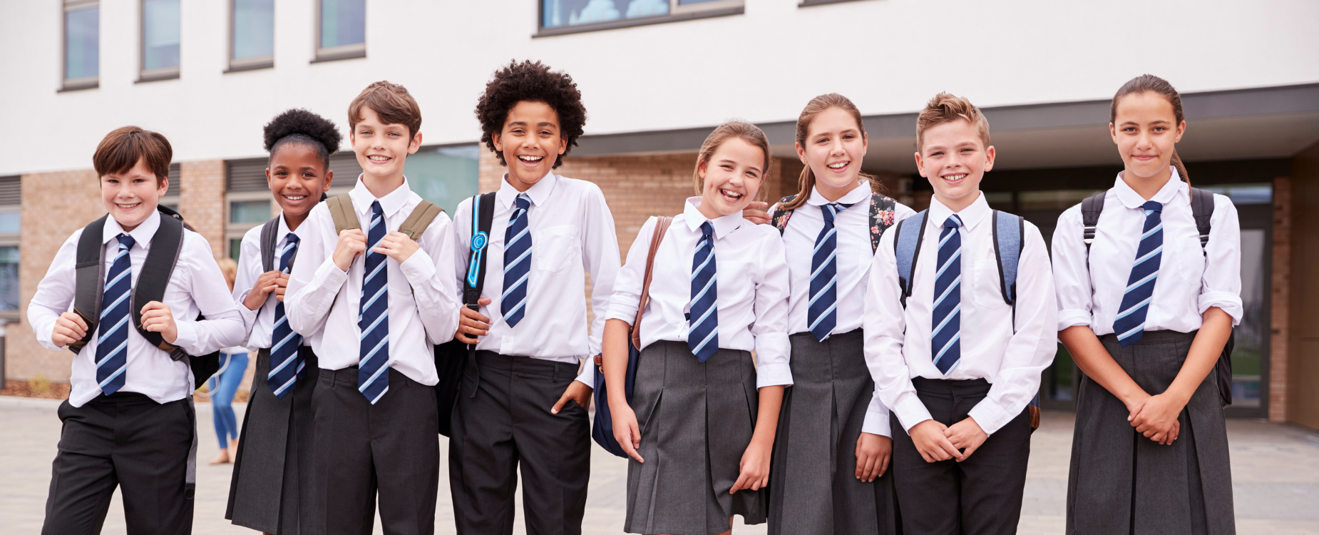 Eight schoolchildren in uniform smiling with backpacks outside a school building.