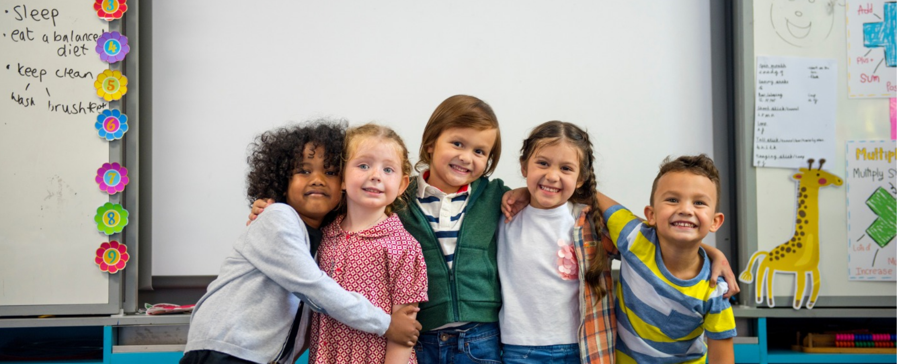 Five smiling young children stand arm in arm in a classroom, posing together in front of a whiteboard.