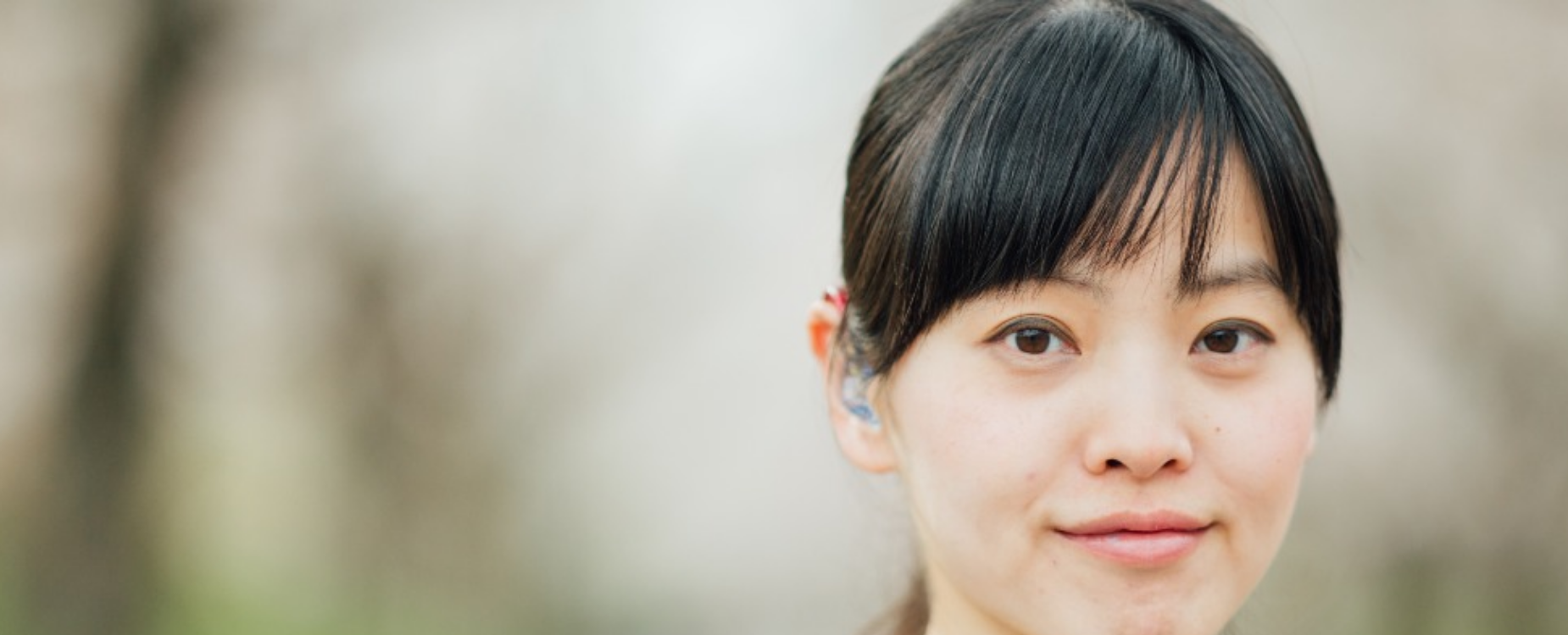 Close-up of a woman wearing a hearing aid, facing the camera outdoors, representing volunteers in the Deaf and hard of hearing community.